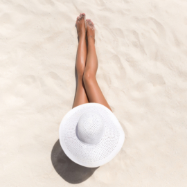 Summer holiday fashion concept - tanning woman wearing sun hat at the beach on a white sand shot from above